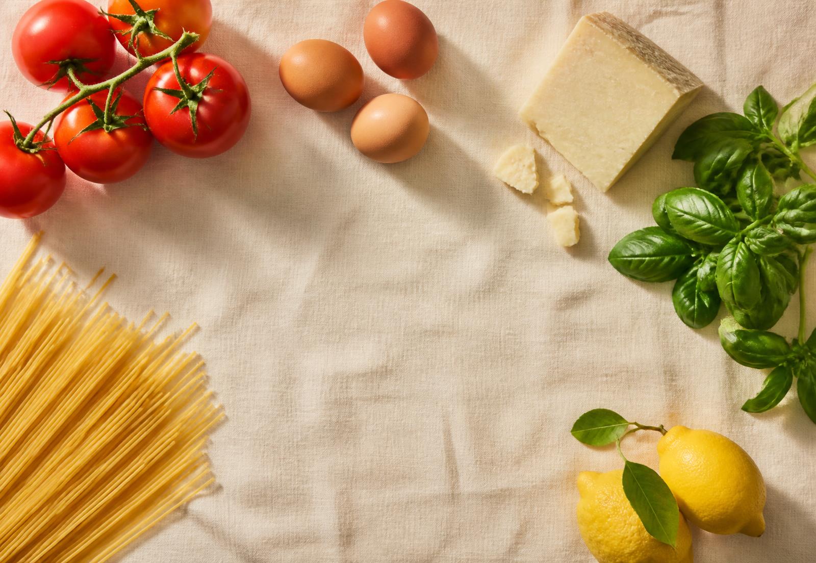 Frische Zutaten wie Tomaten, Eier, Käse, Basilikum und Nudeln auf einem Leinentuch